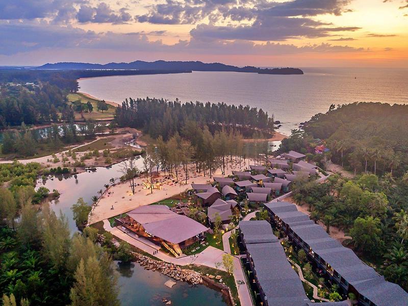 Letecký pohled na resort s bazénem, obklopený lesem a výhledem na moře za západu slunce.