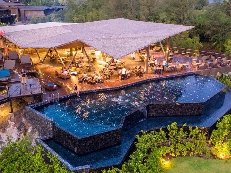 Moderní hotel s osvětleným bazénem a krytou posezením v zeleném prostředí.