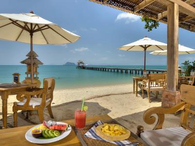 Ristorante sulla spiaggia con tavoli in legno, ombrelloni e vista sul molo e mare turchese.