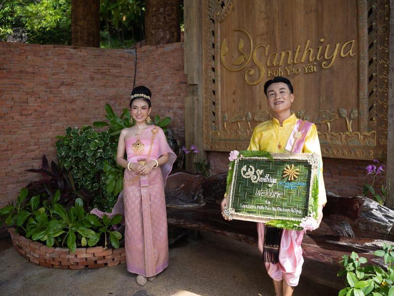 Due persone in abiti tradizionali posano davanti a una parete di legno con piante intorno.
