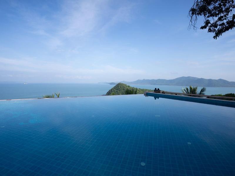 Piscina a sfioro con vista sul mare e colline boschive sotto un cielo sereno.