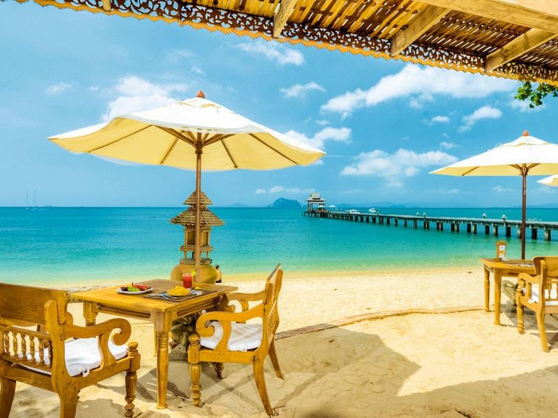 Spiaggia con sedie di legno, tavoli e ombrelloni accanto a un molo sul mare azzurro.