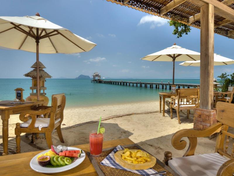 Ristorante sulla spiaggia con tavoli in legno, ombrelloni e vista sul molo e mare turchese.
