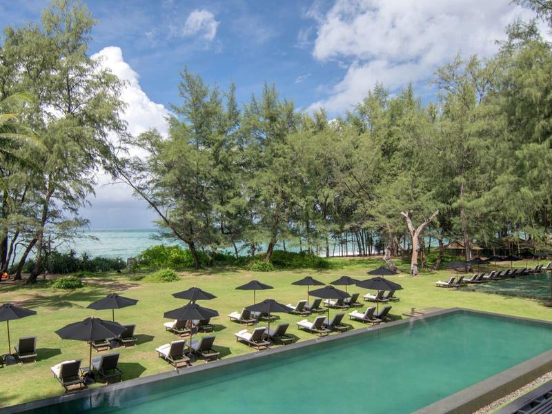 Poolbereich mit Liegestühlen und Schirmen neben grüner Rasenfläche und Bäumen am Meer