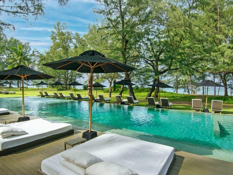 Luxuriöser Pool mit Liegen und Sonnenschirmen umgeben von Bäumen und Blick aufs Meer.