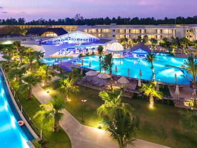 Vue d'un complexe hôtelier moderne avec piscines éclairées et jardins tropicaux au crépuscule.