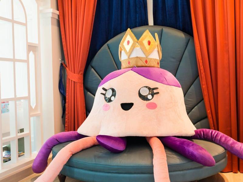 Grand poulpe en peluche rose avec couronne sur chaise élégante devant des rideaux rouges et bleus.