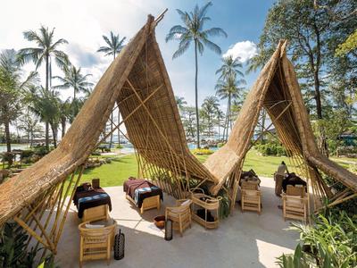 Freiluft-Massagebereich mit Strohdach, Liegestühlen, Palmen und blauem Himmel im tropischen Garten.