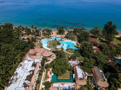 Luftaufnahme eines Resorts mit mehreren Pools, Gebäuden, Bäumen und Meer im Hintergrund.