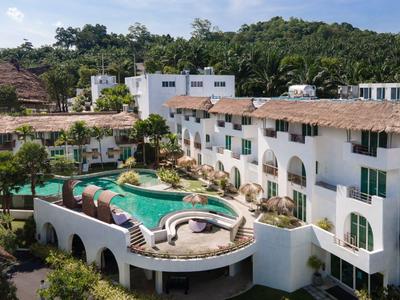 Hotel mit weißer Fassade, rundem Pool auf der Dachterrasse, umgeben von grünen Bäumen und Bergen im Hintergrund.