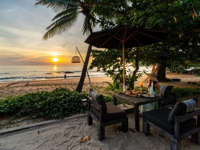 Sonnenuntergang am Strand mit Sitzbereich, Palmen und Segelboot auf dem Meer.