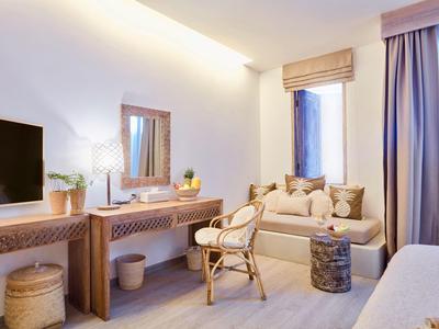 Helles Wohnzimmer mit Holzmöbeln, gemütlichem Sofa, Sessel und großen Fenstern mit Vorhängen.