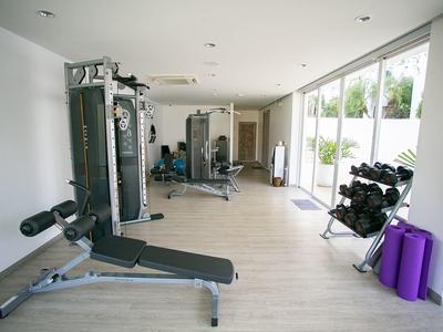 Hell eingerichteter Fitnessraum mit Hantelbank, Kraftstation, Gewichten und Yogamatten am Fenster.