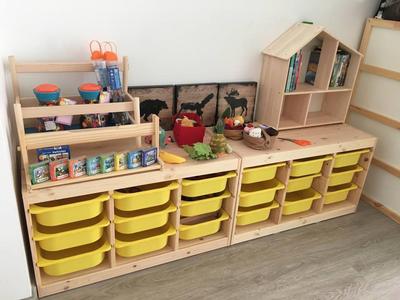 Kindergartenregal mit gelben Plastikboxen, bunten Spielsachen und Puppenhaus aus hellem Holz.