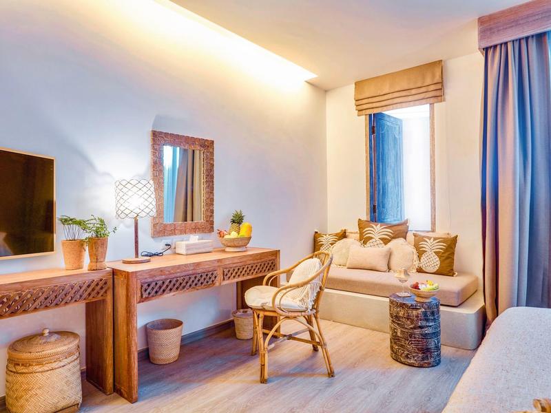 Jasny pokój hotelowy z sofą, biurkiem, krzesłem i dużym oknem z zasłonami.