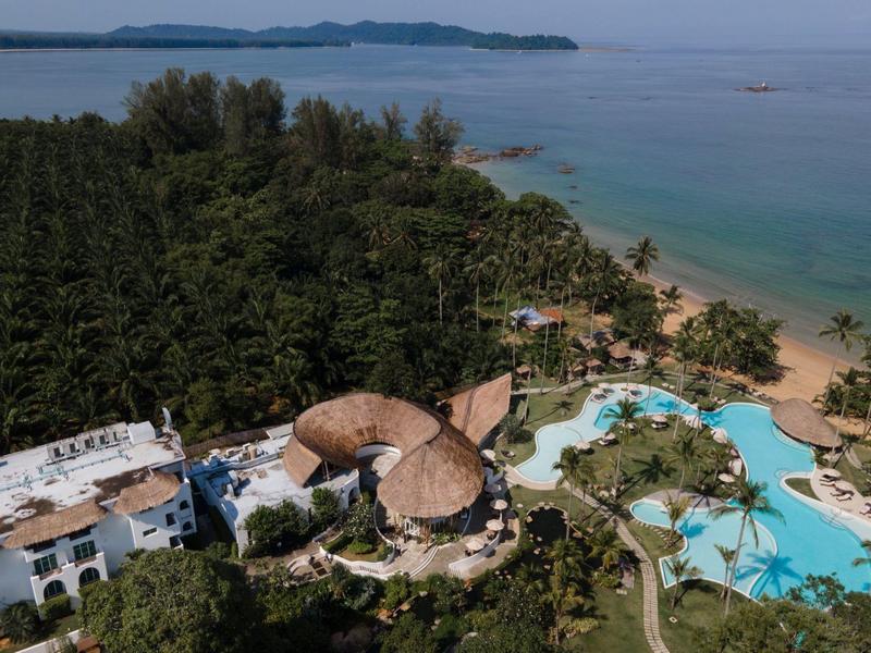 Widok z lotu ptaka na ośrodek wypoczynkowy z basenem, palmami i widokiem na morze i zalesiony półwysep.