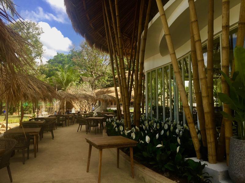 Przestrzeń na zewnątrz hotelu z bambusowymi konstrukcjami, stołami i strzechami pod niebieskim niebem.