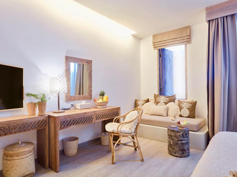 Jasny pokój hotelowy z sofą, krzesłem, telewizorem i biurkiem z lustrem i roślinami.