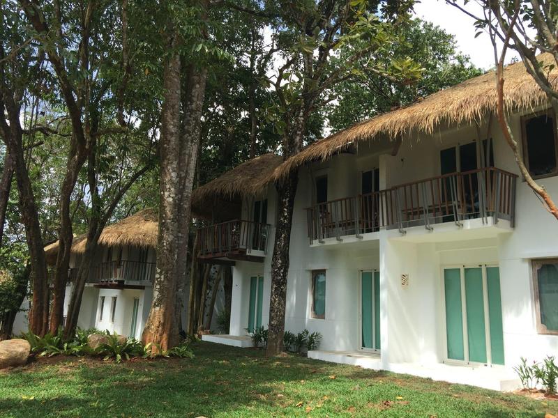 Dwupiętrowy budynek hotelowy z białymi ścianami, zielonymi drzwiami i strzechą otoczony drzewami.