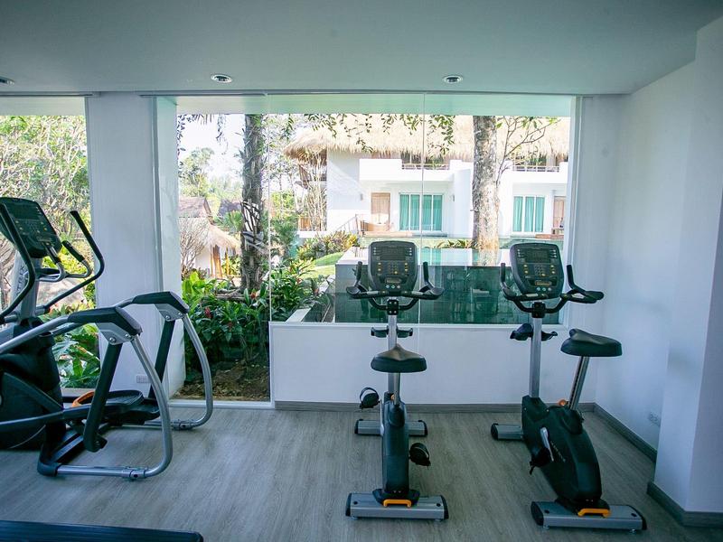 Siłownia z rowerami stacjonarnymi i trenażerami eliptycznymi przed dużymi oknami z widokiem na ogród i budynki.