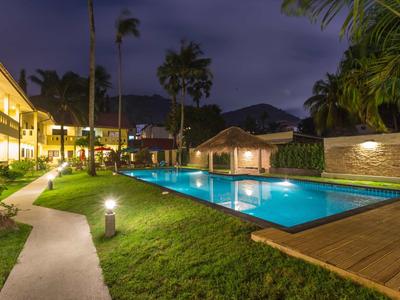 Beleuchteter Außenpool bei Nacht, umgeben von Gras, Palmen und Gebäuden mit warmem Licht.