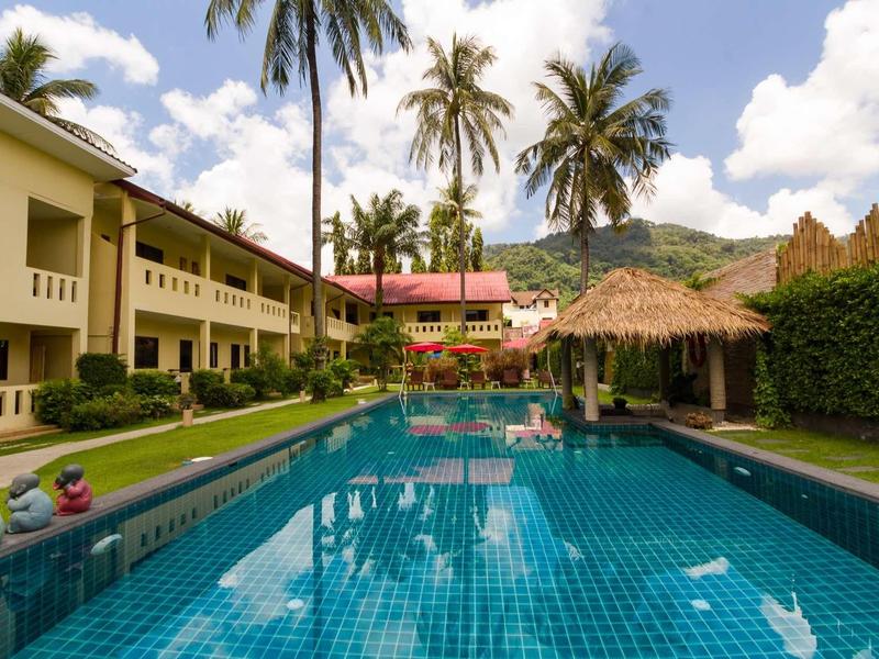Ein blauer Pool umgeben von Gebäuden, Palmen und einer Hütte unter blauem Himmel mit Wolken.
