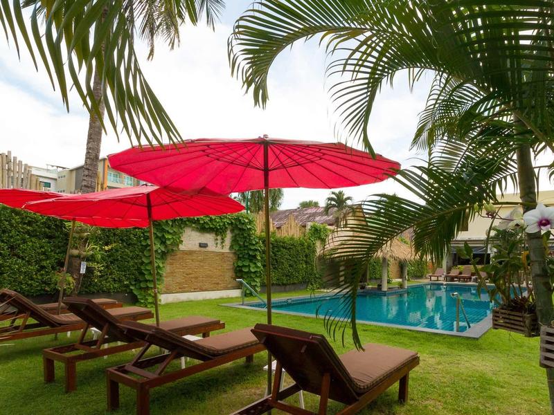 Garten mit grünem Gras, roten Sonnenschirmen, Holzliegen und blauem Pool neben Palmen.