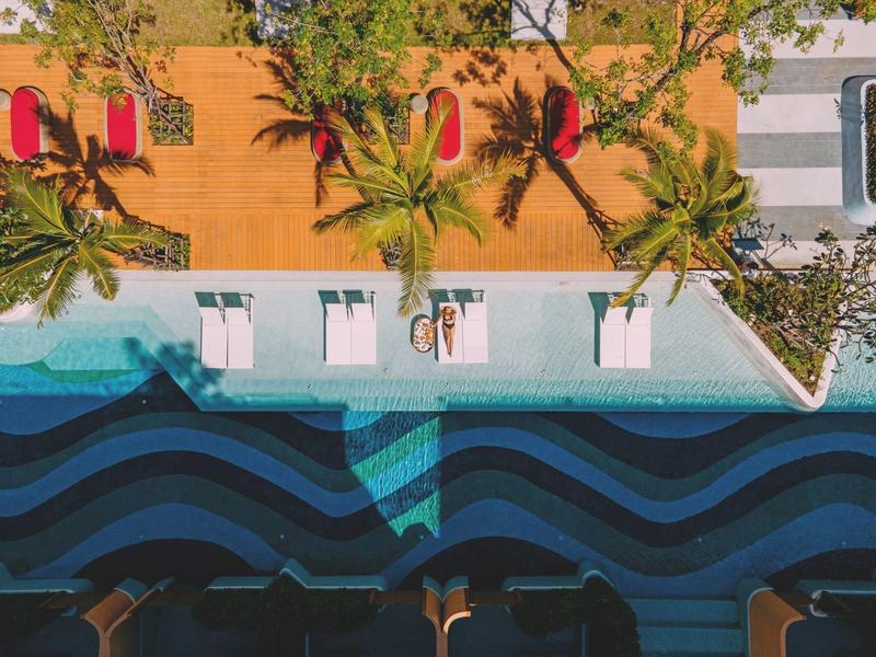 Vista dall'alto di una piscina con motivi ondulati, circondata da lettini e palme.