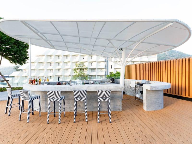 Area bar esterna moderna con tetto rotondo e sgabelli su un deck in legno di fronte a un hotel.