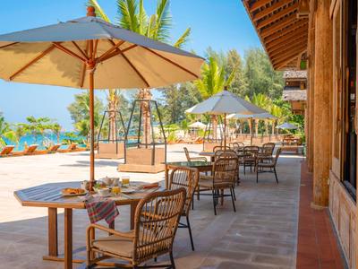 Terras met tafels, stoelen en parasols naast een hotel met uitzicht op tropische palmen.
