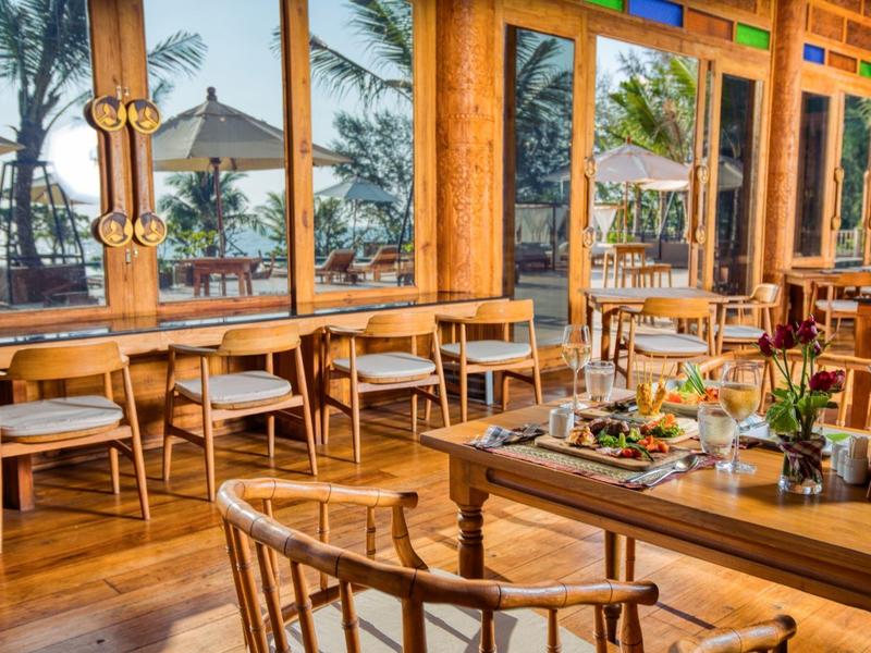 Licht, modern restaurant met houten meubels en grote ramen met uitzicht op palmbomen.