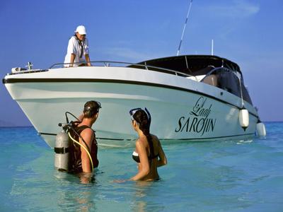 Dva potápěči v mělké vodě vedle lodi pod jasnou oblohou.