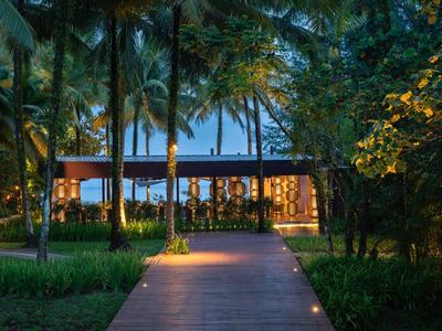 Oświetlona drewniana ścieżka prowadzi do otwartego budynku wśród palm o zmierzchu.