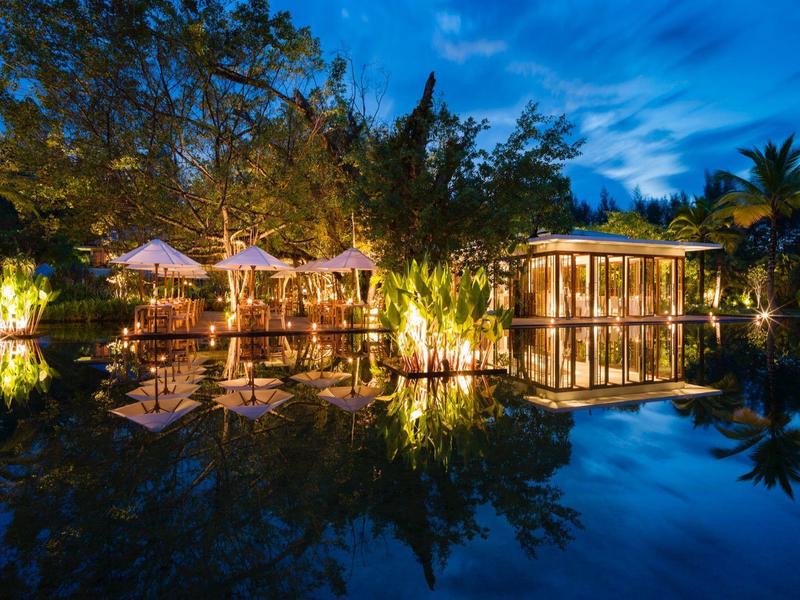 Nocny ogród hotelowy z oświetlonymi pawilonami, drzewami i odbiciem w wodzie pod niebieskim niebem.