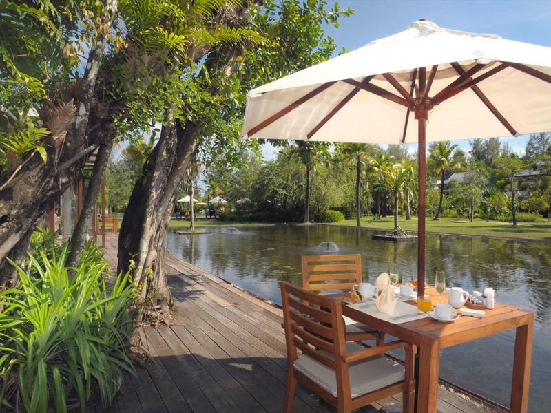 Nakryty stół z dwoma krzesłami i parasolem nad spokojnym brzegiem rzeki otoczonym zielonymi drzewami.