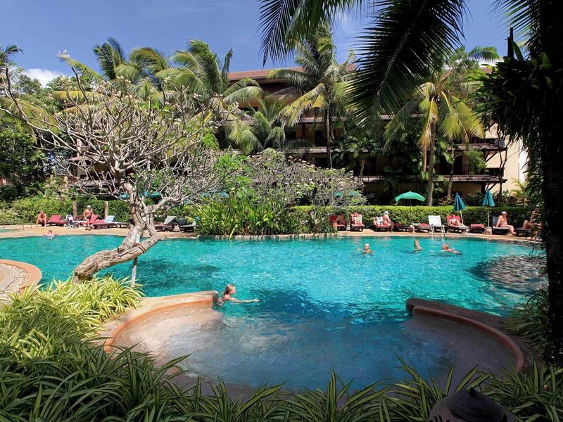 Großer Pool mit klarem Wasser, surrounded von grünen Pflanzen und Palmen unter blauem Himmel.