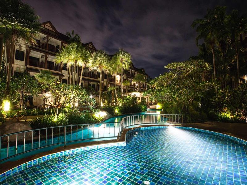 Beleuchteter Pool mit blauen Fliesen umgeben von Palmen und Gebäuden bei Nacht.