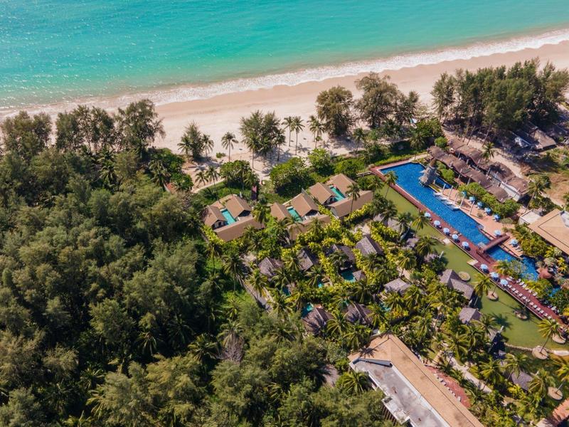 Letecký pohled na hotel s bazénem, palmami a pláží v tropickém prostředí.