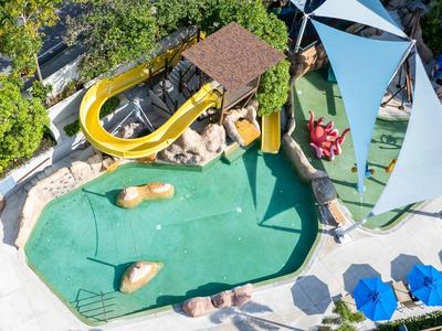 Freizeitbad mit grünem Wasserspielbereich, gelber Rutsche, Sitzbänken und blauen Sonnenschirmen.