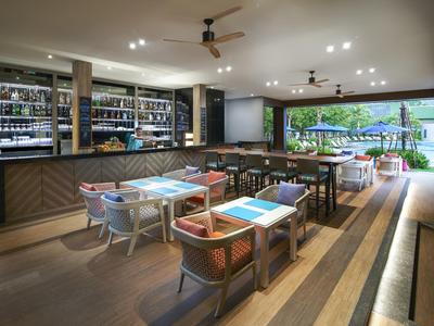 Moderner Gastraum mit Holzboden, farbigen Stühlen, großen Fenstern und einer Bar mit Flaschen.