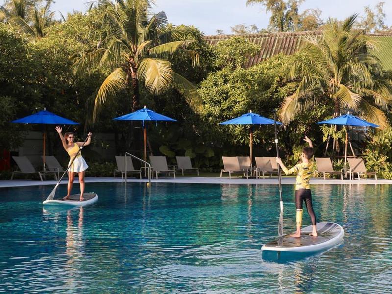 Zwei Personen paddleboarden im Pool, umgeben von Palmen und blauen Sonnenschirmen.