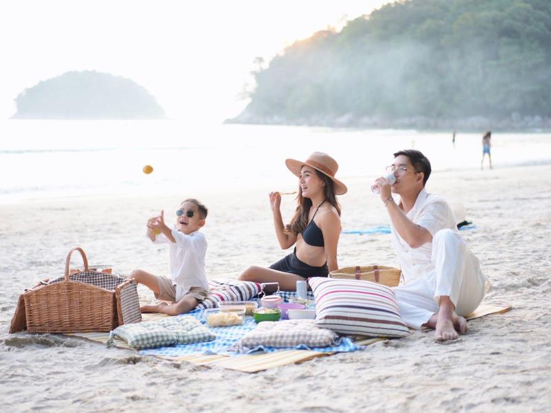 Familie sitzt mit Picknickkorb auf Strandmatte, isst und genießt Sonne am Sandstrand.