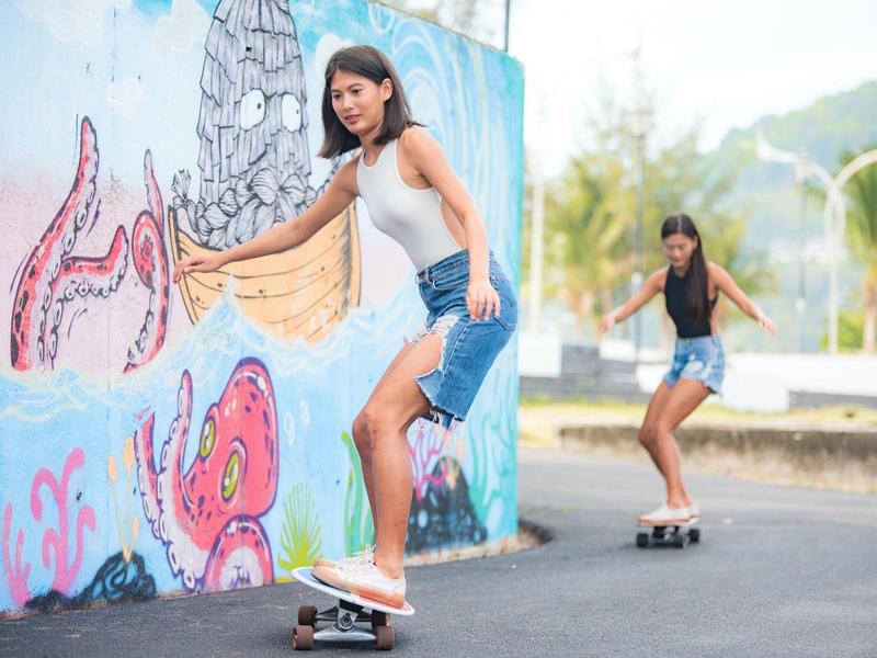 Zwei Mädchen fahren auf Skateboards auf einem Bürgersteig vor einer bunten Wand mit Graffiti.