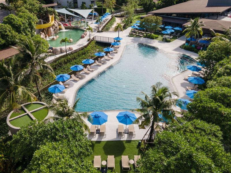 Großer Außenpool mit blauen Sonnenschirmen, grünen Pflanzen und Wasserrutschen in einem Freizeitpark.