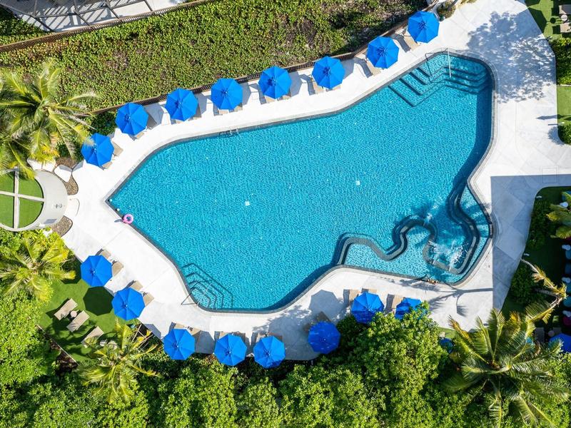 Schwimmbad mit ungewöhnlicher Form, umgeben von blauen Sonnenschirmen, Palmen und grüner Vegetation.