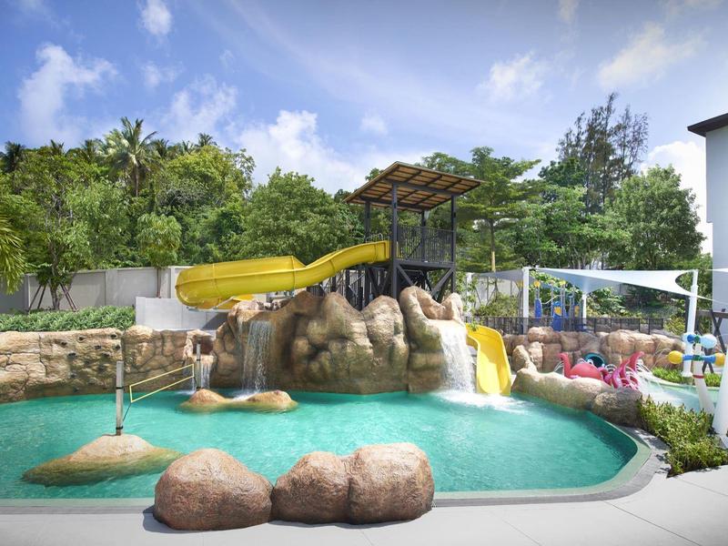 Wasserpark mit Pool, gelber Wasserrutsche, Felsen, grünen Pflanzen und blauem Himmel im Hintergrund.