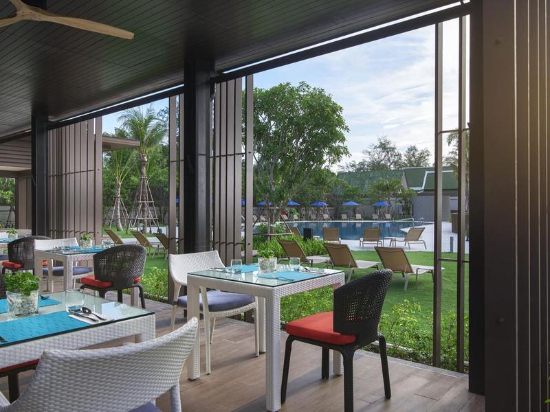 Offenes Restaurant mit Tischen, Stühlen und Blick auf einen Pool und grüne Bäume im Freien.