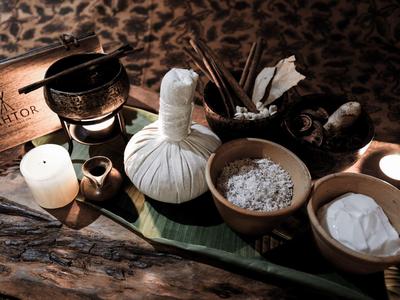 Eine Wellness-Anordnung mit Kräuterpaket, Schalen mit Salz und Pulver, Kerzen und Holzschale auf Holztisch.