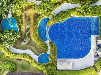 Großer blauer Pool mit runden Formen, umgeben von grünen Sträuchern und geschwungenen weißen Wegen.