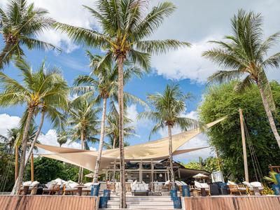 Strandrestaurant mit Palmen, Sonnensegeln und Tischen unter blauem Himmel am Sandstrand.